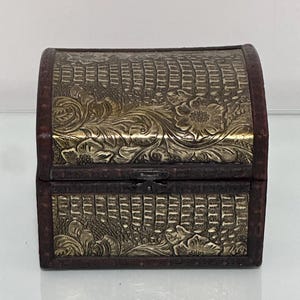 May include: A small rectangular chest with a domed lid, featuring dark brown wood-toned edges and metallic gold-toned panels embossed with a detailed floral pattern and a textured crocodile-skin effect. A small metal latch secures the front.