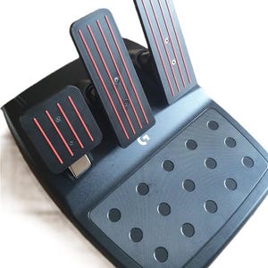 Può includere: Un set di pedali da gioco nero con tre pedali caratterizzati da linee rosse verticali e un poggiapiedi testurizzato con motivi circolari antiscivolo. La base presenta un piccolo logo Logitech G argentato posizionato tra i pedali e il poggiapiedi.