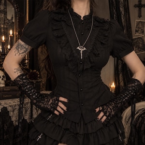 May include: A black short-sleeved blouse featuring a ruffled collar, a button-down front, and a silver bat-shaped pendant necklace. The garment is paired with black lace fingerless gloves that have a corset-style lace-up detail on the forearms.