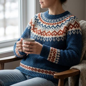 May include: A long-sleeved blue sweater featuring a circular yoke, cuffs, and hem decorated with a geometric pattern in cream, light gray, and rust orange. The sweater has a ribbed crew neckline, ribbed cuffs, and a ribbed hem, with a textured knit.