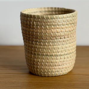May include: A cylindrical woven basket crafted from light tan and pale green dried grass fibers. The container features a consistent, tight coil weave pattern throughout its exterior, creating a textured surface with a wide, open top.