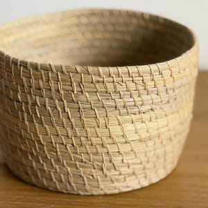 May include: A round, open-top basket constructed from light tan, woven natural fibers. The basket features a consistent, tight weave pattern throughout its cylindrical body, creating a textured surface with a neutral, earthy color palette.