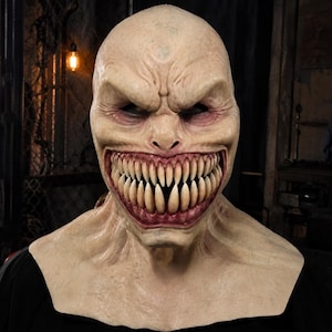 Op de afbeelding: Een masker voor het hele hoofd met een bleke, huidkleurige teint en een gladde, haarloze schedel. Het gezicht heeft diepliggende, donkere ogen en een wijd opengesperde mond vol met meerdere rijen scherpe, gebroken witte tanden.