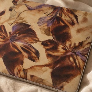 Könnte beinhalten: Eine Laptop-Hülle mit einem Blumenmuster im Aquarellstil, das große dunkelbraune und tiefviolette Blütenblätter auf einem hellbeigen, pergamentartigen Hintergrund zeigt und die flache, rechteckige Oberfläche bedeckt.