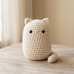May include: A cream-colored crochet cat plush toy with a rounded body, two small pointed ears, and a curved tail. The cat features two round black plastic eyes and a small pink stitched nose, all crafted with a consistent textured yarn stitch.