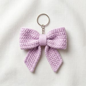 May include: A light purple crochet bow keyring featuring a textured yarn construction with two loops and two hanging tails. The bow is secured at the centre with a matching yarn wrap and is attached to a circular silver-toned metal split ring.