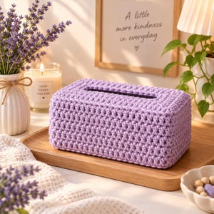 Puede incluir: Una funda rectangular para caja de pañuelos hecha de hilo morado claro con un patrón de punto de ganchillo denso. La funda presenta una abertura alargada centrada en la parte superior para dispensar pañuelos, con textura.