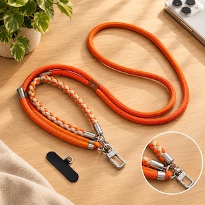 May include: A bright orange rope phone lanyard featuring a braided beige and orange accent cord, silver-toned metal hardware, and a black rectangular phone tether tab. The lanyard includes a long adjustable strap and a shorter wrist loop attachment.