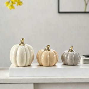 May include: A set of three decorative ceramic pumpkins in varying sizes, featuring a smooth, glossy finish in cream, light beige, and soft grey. Each pumpkin is topped with a polished, reflective gold-coloured stem, creating a distinct contrast.