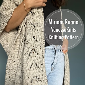 May include: A beige knitted shawl with a lacy pattern and speckles of brown yarn. The shawl is draped over a person's shoulders and is being held in place by one hand. The text "Miriam Ruana VanessaKnits Knitting Pattern" is visible in the image.