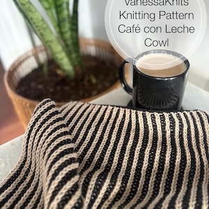 Puede incluir: Un cuello a rayas negras y beige, parte de la colección VanessaKnits Knitting Pattern Café con Leche, está drapeado cerca de una taza negra llena de café. Una planta en maceta está en el fondo.