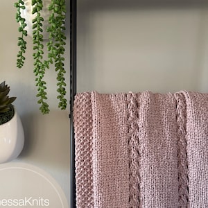 Op de afbeelding: Een roze gebreide deken met een kabelsteekpatroon. De deken is gedrapeerd over een zwart metalen plank met een houten bovenkant. De tekst "VanessaKnits Knitting Pattern Signs of Spring" is zichtbaar in de afbeelding.