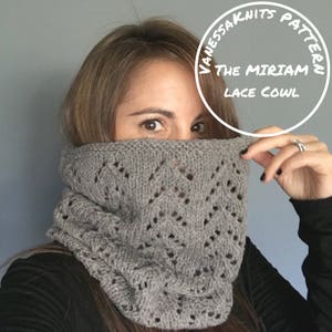 Op de afbeelding: Een grijze gebreide sjaal met een kantpatroon. De sjaal wordt gedragen door een persoon, die gedeeltelijk hun gezicht bedekt. De tekst "Vanessa Knits Pattern The Miriam Lace Cowl" is zichtbaar in een cirkel boven het hoofd van de persoon.