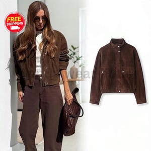 May include: A dark brown suede jacket featuring a high collar, a front zipper closure with a snap button placket, and two front pockets with snap button flaps. The jacket has long sleeves and a straight hemline with a subtle elasticated finish.
