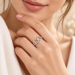 Puede incluir: Un anillo de tono plateado pulido que presenta un diseño de símbolo de infinito formado por dos corazones interconectados. La banda es lisa y metálica, mostrando una estructura de bucle continuo que descansa sobre el dedo con un acabado limpio y brillante.