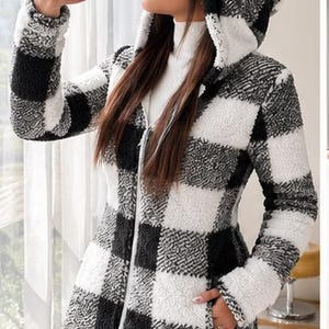 May include: A long-sleeved hooded jacket featuring a black and white buffalo check pattern. The garment is constructed from a textured, fuzzy fleece material and includes a full-length front zip closure, a hood, and two side pockets.