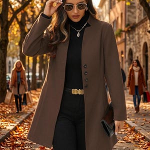 May include: A brown long-sleeved coat with a stand-up collar and a single row of four dark buttons down the front. The coat is worn over a black turtleneck sweater and black trousers, accessorized with a gold-toned belt and a black clutch.