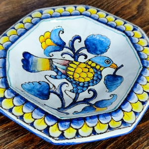Könnte beinhalten: Eine achteckige Keramikschale mit weißem Grund, die einen handbemalten blau-gelben Vogel zeigt, der zwischen stilisierten Blumenranken sitzt. Der Rand ist mit einem wiederkehrenden Muster aus blau-gelben Blütenblättern verziert.