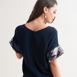 Puede incluir: Top azul marino con mangas de lentejuelas. El top tiene cuello en V y mangas cortas con lentejuelas plateadas y negras. La modelo lleva un collar y pendientes dorados.