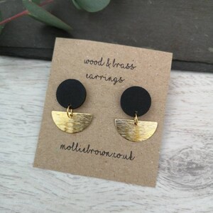 May include: A pair of black and gold earrings. The earrings are made of wood and brass. The black circles are made of wood and the gold crescent shapes are made of brass. The earrings are displayed on a brown card with the text "wood & brass earrings" and "molliebrown.co.uk".