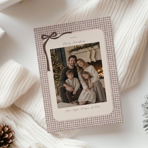 May include: A rectangular Christmas card featuring a beige and white gingham border with a dark brown illustrated ribbon bow. The center displays a photograph of a family with the text 2026, Merry Christmas, and John, Taylor, Morgan, & Nora.
