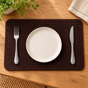 May include: A rectangular dark brown crocheted placemat featuring a dense, textured stitch pattern. Centred on the placemat is a plain white round ceramic plate, flanked by a stainless steel fork on the left and a stainless steel knife on the right.