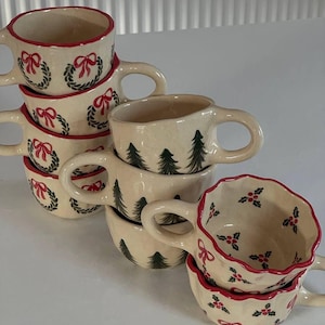Könnte beinhalten: Eine Sammlung cremefarbener Keramiktassen mit handgemalten weihnachtlichen Motiven. Einige Tassen zeigen grüne Kränze mit roten Schleifen, andere dunkelgrüne Tannenbäume und einige rote Stechpalmenbeeren mit grünen Blättern und gewelltem Rand.
