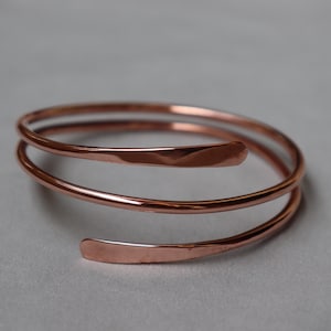 Può includere: Un bracciale in rame lucido caratterizzato da un design a spirale continua con tre spire sovrapposte. Il metallo ha una calda tonalità bruno-rossastra metallica e una superficie liscia e riflettente con estremità leggermente appiattite.