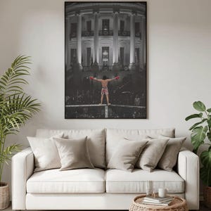 Pode incluir: Uma impressão de arte de parede emoldurada em preto e branco apresentando um boxeador em pé na borda de um ringue com os braços estendidos, sobreposto ao exterior da Casa Branca. O boxeador usa luvas vermelhas e shorts estampados em uma arena.
