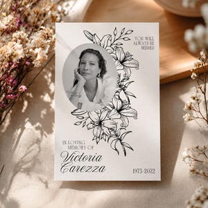 Könnte beinhalten: Eine rechteckige Gedenkkarte mit einem schwarz-weißen ovalen Porträt einer Person, umgeben von schwarzen Strichzeichnungen von Lilien. Der Text lautet: You will always be missed, In loving memory of Victoria Carezza, und 1975-2022.