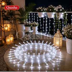 Pode incluir: Uma bobina de luzes LED transparentes que emitem um brilho branco intenso, disposta sobre uma mesa redonda. As luzes possuem numerosas lâmpadas pequenas espaçadas uniformemente dentro de um tubo flexível e transparente.