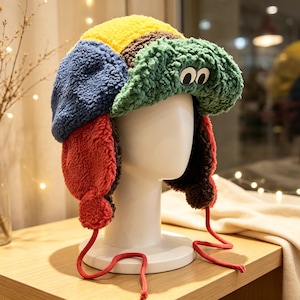 May include: A multi-colored plush trapper hat featuring a yellow crown, blue side panels, and a green front brim with two white and black eye appliqués. The hat includes red ear flaps with matching red ties and a dark brown interior lining.