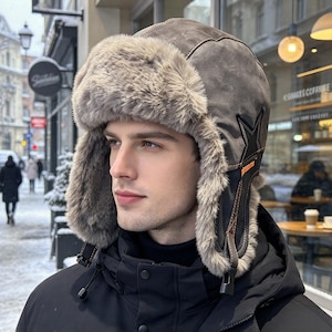 May include: A grey trapper hat featuring a soft, light brown faux fur lining and trim. The exterior is made of a smooth, matte grey material with dark grey star-shaped stitching on the side and orange accent stitching near the adjustable chin strap.
