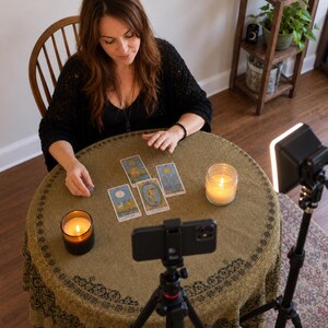 May include: A round table covered with an olive green cloth featuring a black border pattern, displaying four tarot cards arranged in a spread. Two lit candles in glass jars sit on the table alongside a smartphone mounted on a tripod for recording.