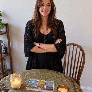 May include: A person with long brown hair wearing a black top and a black crocheted cardigan, standing behind a round table. On the table are three tarot cards, two lit candles in glass jars, and a small wooden bowl containing various crystals.