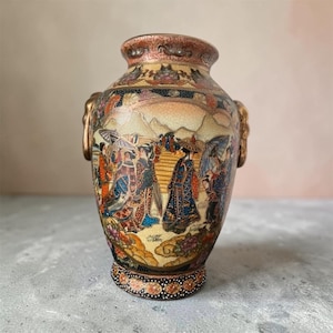 以下が含まれることがあります： 2つの金色の取っ手が付いた陶器製の花瓶。伝統的な衣装を着て傘を差した人物が描かれた詳細な絵柄が特徴です。表面はひび割れたような質感で、金、青、赤、黒のアクセントが施され、縁には装飾的な模様が描かれています。