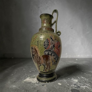 Puede incluir: Una jarra de cerámica con acabado verde texturizado, que muestra un caballo marrón oscuro y una figura en un carro en rojo y negro. El recipiente tiene un asa, un cuello estrecho con patrones decorativos y una base circular negra sólida.