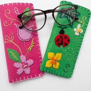 Könnte beinhalten: Zwei rechteckige Brillenetuis aus Filz, eines in Pink und eines in Grün, mit handgestickten Blumen- und Insektenmotiven sowie Perlenverzierungen. Das pinke Etui zeigt ein Blumen- und Rankendesign, während das grüne Etui einen Marienkäfer und Blüten aufweist. Eine Brille mit Schildpattmuster liegt darauf.