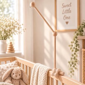 May include: A light-colored wooden crib mobile arm featuring a vertical post, an angled extension arm, and a small metal hook at the end. The arm is secured to the crib rail with two cloud-shaped wooden clamps, showcasing a smooth, natural finish.