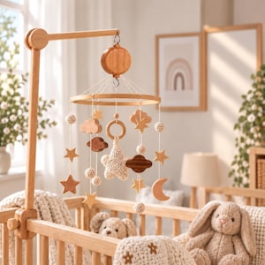 Pode incluir: Um móbile de madeira para bebé com ornamentos suspensos, incluindo estrelas, nuvens, uma lua crescente e uma estrela central de croché. O móbile está suspenso num braço de madeira clara e inclui várias esferas texturizadas.