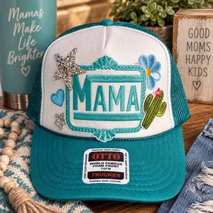 May include: A teal and white trucker hat featuring a central teal rectangular frame with the word MAMA in teal lettering. The hat is decorated with a rhinestone star, a small blue heart, a blue flower, and a green cactus patch. An Otto brand sticker is on the brim.