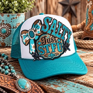 May include: A white and teal trucker hat featuring an embroidered patch with a turquoise bow, brown accents, and the text SALTY Just STFU in bold teal lettering. The cap includes dark blue star-shaped embellishments and a mesh back panel.
