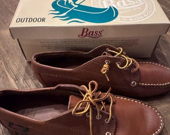 May include: A pair of brown leather boat shoes with yellow and brown laces, featuring white contrast stitching and a small Bass logo tag. The shoes are positioned in front of a rectangular cardboard box labeled Bass Outdoor with a canoe graphic.