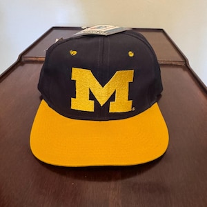 Puede incluir: Una gorra de béisbol azul marino con una letra M dorada grande bordada en el panel frontal. La gorra tiene una visera plana de color dorado sólido e incluye una pequeña etiqueta adherida en la parte superior con la palabra Universal.