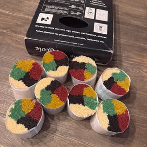 May include: A black Nicole Creations latch hook kit box with instructions and eight cylindrical bundles of yarn. Each yarn bundle features a multi-colored pattern of red, yellow, green, black, and cream segments wrapped in white paper bands.