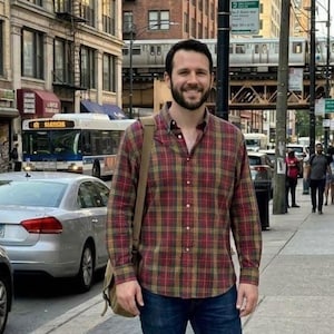 May include: A long-sleeved button-down shirt featuring a red, yellow, and green plaid pattern, worn with dark blue denim jeans and a tan shoulder bag, standing on a city sidewalk with a bus and elevated train in the background.