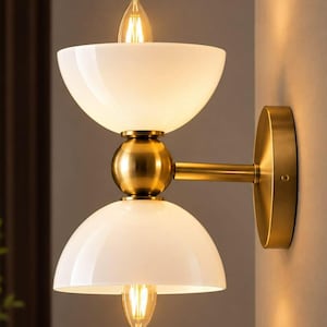 Könnte beinhalten: Eine Wandleuchte mit gebürstetem Messing-Finish und zwei weißen, schalenförmigen Glasschirmen. Das Design umfasst eine zentrale Messingkugel, die die beiden Schirme verbindet, wobei jeder Schirm eine leuchtende Filament-Glühbirne enthält.