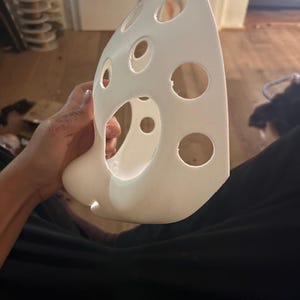 Könnte beinhalten: Eine weiße, geschwungene, starre Maske mit einer Reihe von kreisförmigen Ausschnitten unterschiedlicher Größe auf der Oberfläche. Die Maske hat eine glatte, matte Oberfläche und eine spitze, längliche Form, die sich nach oben hin verjüngt.