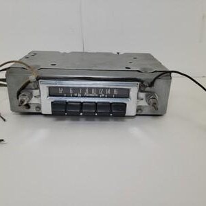 May include: A vintage automotive radio unit featuring a rectangular silver metal chassis, a central display panel with AM frequency markings, and five black rectangular push buttons. The device includes two metal control knobs and loose wires.