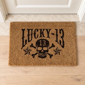 Könnte beinhalten: Eine rechteckige, hellbraune Kokosfasermatte mit schwarzem Grafikdruck. Das Design zeigt den Schriftzug LUCKY-13 oben, ein zentrales Totenkopf-Symbol mit gekreuzten Knochen und der Zahl 13 auf der Stirn sowie einen fünfzackigen Stern.